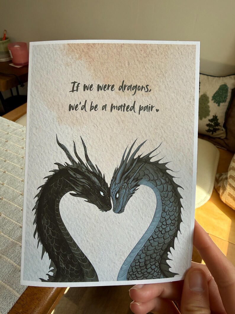 Op de afbeelding: Wenskaart met twee draken die een hart vormen. E&eacute;n draak is zwart, de andere blauwgrijs. De tekst luidt: 'Als we draken waren, zouden we een paar zijn.' De kaart heeft een achtergrond in aquarel-stijl.