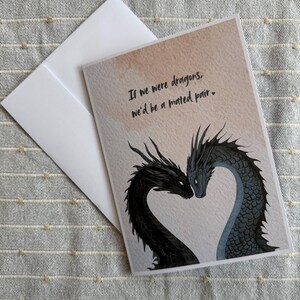Op de afbeelding: Wenskaart met een drakenillustratie. Twee draken, een zwarte en een blauwe, vormen een hart. De kaart bevat de tekst: "If we were dragons, we'd be a mated pair." Een witte envelop is inbegrepen.