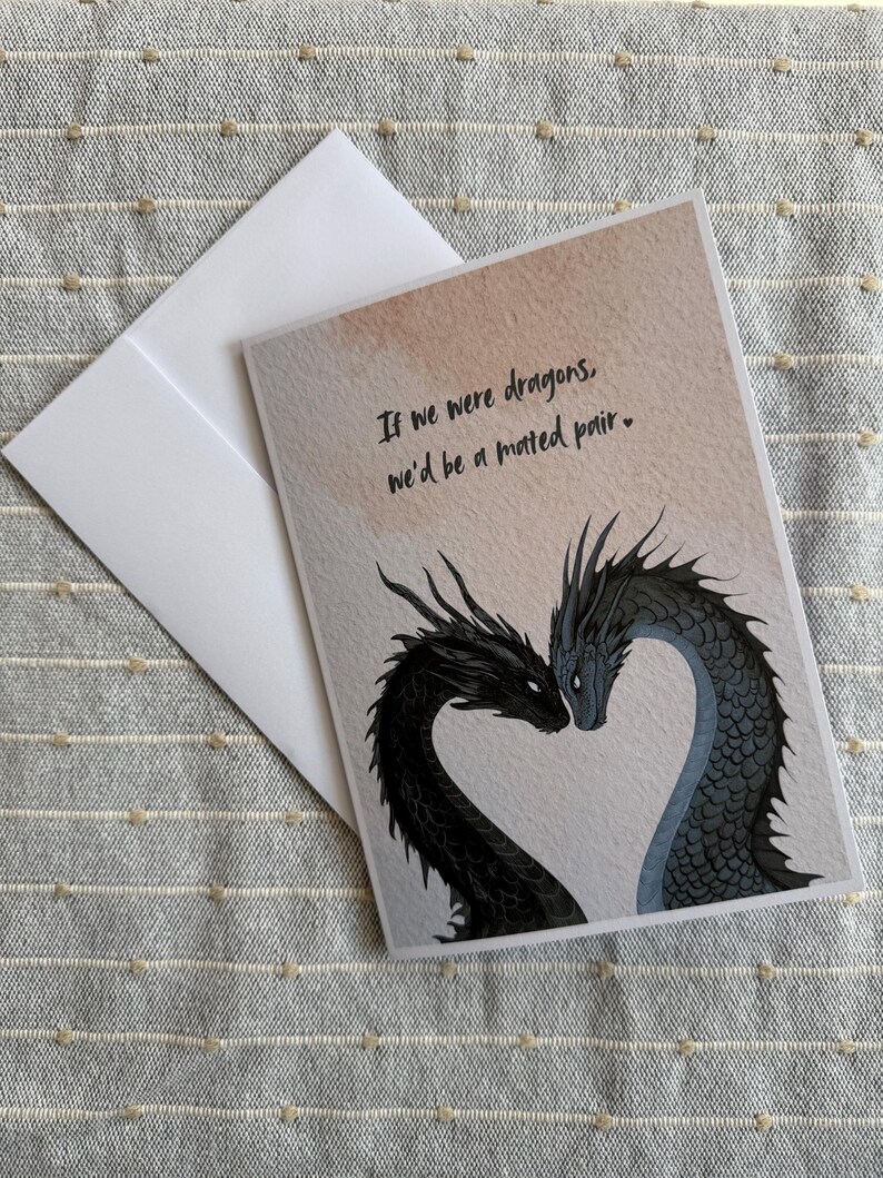 Op de afbeelding: Wenskaart met een drakenillustratie. Twee draken, een zwarte en een blauwe, vormen een hart. De kaart bevat de tekst: "If we were dragons, we'd be a mated pair." Een witte envelop is inbegrepen.
