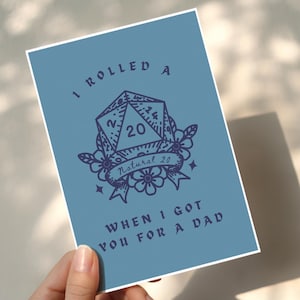 May include: A blue card with the text "I rolled a natural 20 when I got you for a dad." The card features a die with the number 20, surrounded by floral elements. The card is held by a hand.