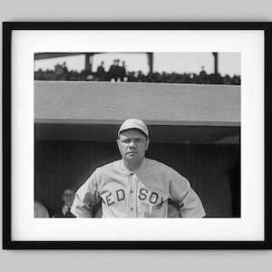 May include: A black and white photo of a baseball player wearing a white cap and a jersey with the text "RED SOX" on the front. The player is standing in a baseball dugout with a crowd of people in the background.