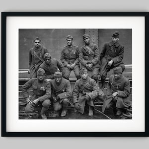 May include: Black and white photograph of eight men in military uniform. The men are standing and sitting on a platform. They are all looking at the camera. The men are wearing hats and have medals on their uniforms.