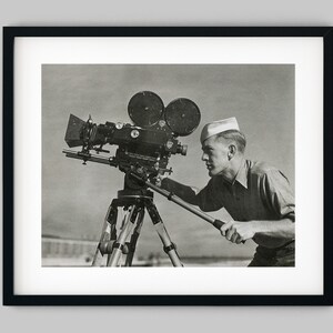 May include: A black and white photograph of a person wearing a white sailor hat operating a large film camera on a tripod.