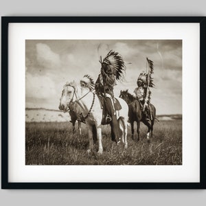 Könnte beinhalten: Zwei Indianer zu Pferd, die traditionelle Kopfbedeckungen und Kleidung tragen. Die Männer reiten weiße Pferde auf einer grasbewachsenen Weide.