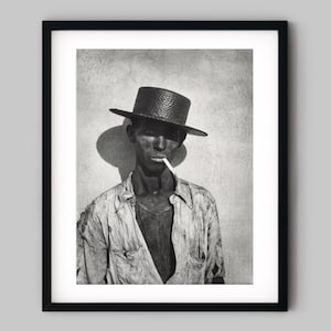 May include: Black and white framed photograph of a person wearing a straw hat and a button-down shirt, with a cigarette in their mouth. The image is a portrait with a shadow cast on the wall behind the person.