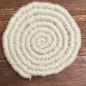 Puede incluir: Un posavasos redondo verde claro hecho de hilo enrollado. El posavasos tiene un diseño en espiral, con el hilo cuidadosamente enrollado desde el centro hacia afuera. El hilo tiene una textura suave y difusa, y las puntadas son visibles.