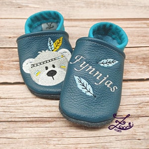 K&ouml;nnte beinhalten: Ein Paar babyblaue Lederschuhe mit einem wei&szlig;en B&auml;ren, der ein Stirnband mit Federn tr&auml;gt, das auf einem Schuh gestickt ist, und dem Namen "Fynnjas", der auf dem anderen Schuh gestickt ist.