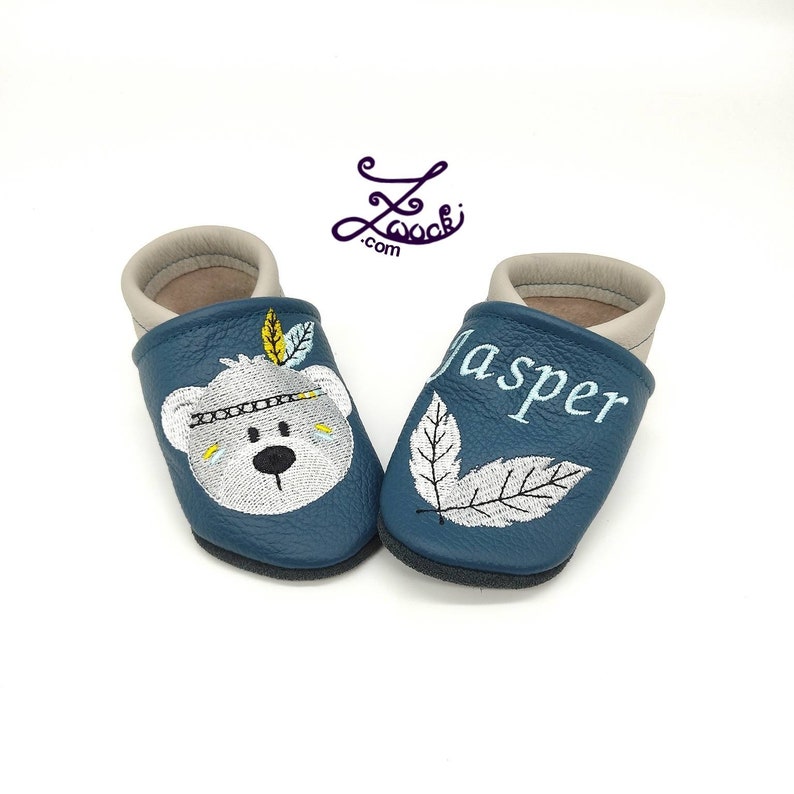 K&ouml;nnte beinhalten: Ein Paar blaue Babyschuhe mit gestickten Designs. Ein Schuh zeigt einen wei&szlig;en B&auml;ren mit einem bunten Kopfschmuck und der andere Schuh hat den Namen "Jasper" gestickt.