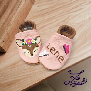 Könnte beinhalten: Rosa Lederbabyschuhe mit gestickten Hirsch- und Blumendesigns auf einem Schuh und dem Namen "Lene" und einem Schmetterling auf dem anderen Schuh.