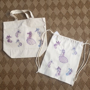 May include: Two canvas tote bags with a seahorse design. The larger bag has a top handle and the smaller bag has drawstrings. Both bags are white with purple and blue seahorses.