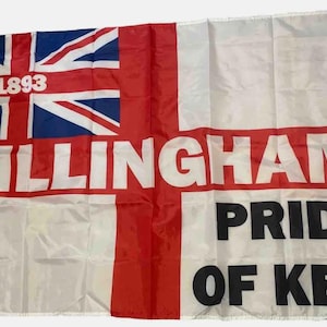 May include: A white flag with a red cross and a Union Jack in the top left corner. The words "Gillingham Pride of Kent" are printed in black letters on the flag. The year "1893" is printed in white letters on the Union Jack.
