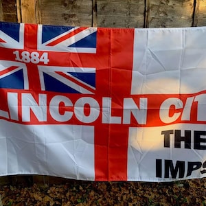 Może przedstawiać: Biała flaga z czerwonym krzyżem i Union Jack w lewym górnym rogu. Flaga zawiera napis "LINCOLN CITY THE IMPS" czarnymi literami i rok "1884" czerwonymi literami.