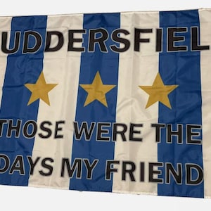 Puede incluir: Una bandera a rayas blancas y azules con las palabras "Huddersfield" y "Those were the days my friend" impresas en negro. Tres estrellas doradas están centradas en la bandera.
