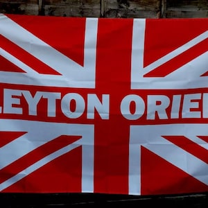 Könnte beinhalten: Eine rot-weiß karierte Flagge mit dem Union Jack-Design und den weißen Worten "LEYTON ORIENT". Die rechteckige Flagge besteht aus Stoff, wahrscheinlich zur Präsentation.