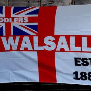 May include: A white flag with a red cross and a blue Union Jack in the top left corner. The words "SADDLERS" and "WALSALL" are printed in white on the flag. The words "EST. 1888" are printed in black in the bottom right corner.