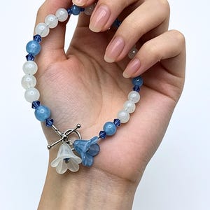 May include: A beaded bracelet featuring alternating white and light blue round beads, accented with small dark blue crystal spacers. The bracelet includes a silver-toned toggle clasp with two hanging flower-shaped charms in white and blue.