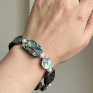 May include: A bracelet with iridescent abalone shell pieces and white pearls. The abalone shell has a mix of green, blue, and purple hues. The bracelet is strung together with a black cord.