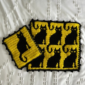 May include: Two crocheted cat-themed squares. One square features a single black cat silhouette on a yellow background. The other square has a larger design with multiple black cat silhouettes on a yellow background. Both have a black border.