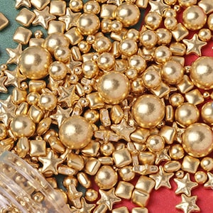 Puede incluir: Primer plano de una variedad de espolvoreados de color dorado, incluyendo estrellas, cuadrados y bolas redondas. Los espolvoreados están esparcidos sobre una superficie roja.