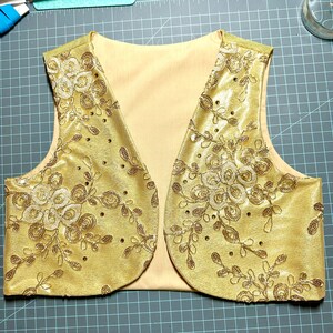 May include: A gold vest with a floral pattern and sequins. The vest has a V-neck and is sleeveless.