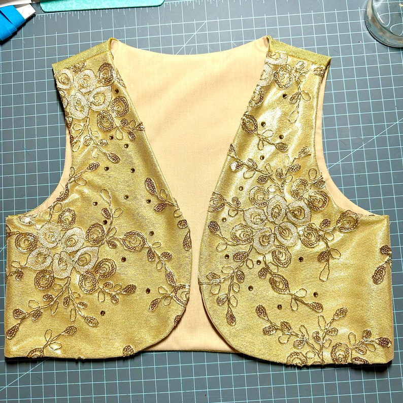 May include: A gold vest with a floral pattern and sequins. The vest has a V-neck and is sleeveless.