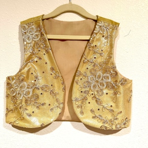 May include: A gold vest with a V-neck and intricate floral embroidery. The vest is made of a shiny fabric and has a cropped length.