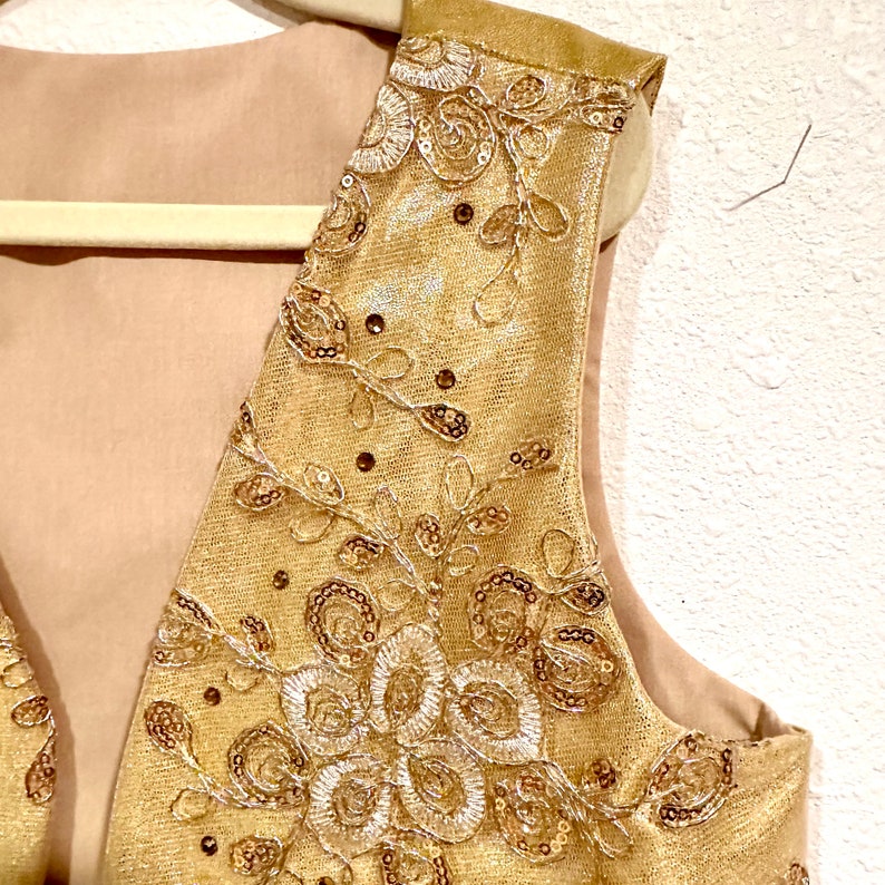 May include: A gold sleeveless vest with a delicate floral pattern and sequins. The vest is made of a sheer fabric and has a V-neckline.