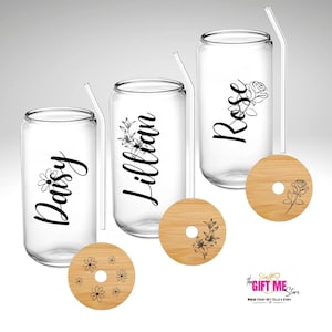 May include: Three clear glass can-shaped cups with black straws and personalized names: Daisy, Lillian, and Rose. Each cup has a matching bamboo lid with a floral design.