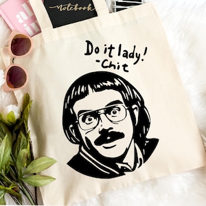 May include: A beige canvas tote bag with a black graphic of a man with glasses and a mustache, and the text "Do it lady! -Chit". The bag contains a pink notebook, a black notebook, a phone, and sunglasses. The bag has a pair of pink shoes and flowers.
