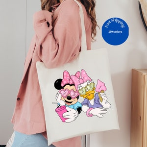 Op de afbeelding: Een witte canvas tas met een kleurrijke afbeelding van Minnie Mouse en Daisy Duck met hartvormige zonnebrillen. Minnie houdt een roze drankje vast en Daisy een roze ijsje. Beide figuren dragen roze strikken op hun hoofd.