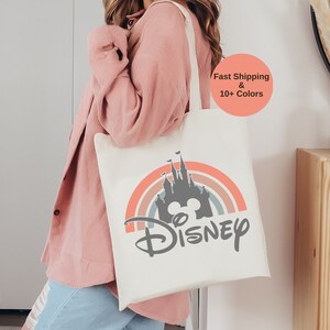 K&ouml;nnte beinhalten: Eine wei&szlig;e Canvas-Tragetasche mit einem Regenbogen und einer Silhouette von Schloss Aschenputtel mit dem Wort "DISNEY" in grauer Schrift.