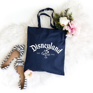 May include: A navy blue canvas tote bag featuring the white text Disneyland EST. 1955 with a line art illustration of Mickey Mouse. The bag has two shoulder straps and is accompanied by a pair of blue and white striped flats and floral accents.