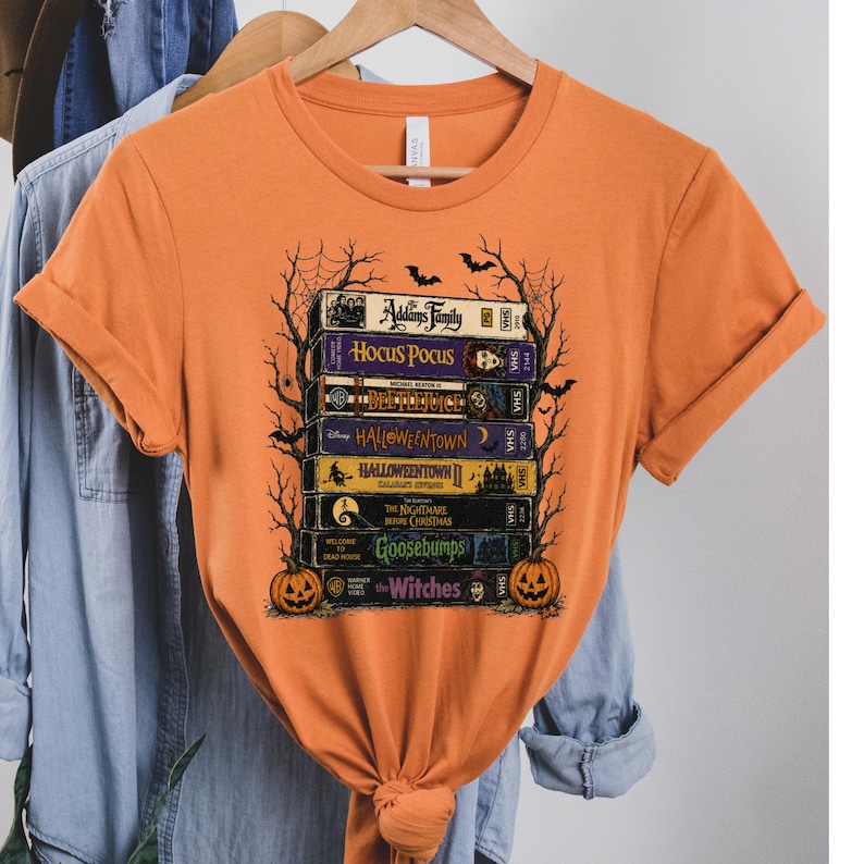 Puede incluir: Una camiseta naranja de manga corta con un estampado gr&aacute;fico de una pila de cintas VHS tituladas The Addams Family, Hocus Pocus, Beetlejuice, Halloweentown, Halloweentown II, The Nightmare Before Christmas, Goosebumps y The Witches con calabazas.