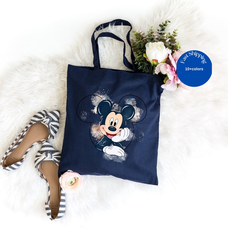 May include: A navy blue tote bag featuring a graphic print of Mickey Mouse centered within a circular outline, surrounded by a watercolor-style splash effect. The bag has two matching navy blue fabric handles attached to the top edge.