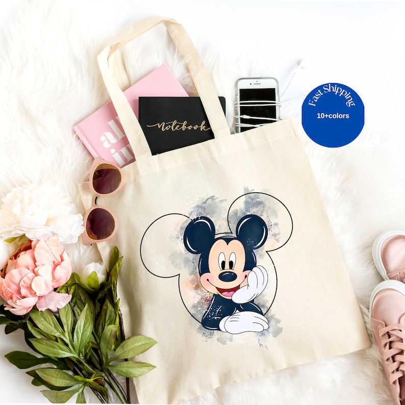 May include: A beige canvas tote bag featuring a graphic of Mickey Mouse inside a circular outline. The bag contains a pink book, a black notebook with the word Notebook printed on the cover, and a smartphone with white wired earbuds.