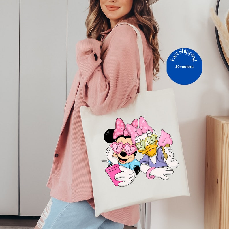 Op de afbeelding: Een witte canvas tas met een kleurrijke afbeelding van Minnie Mouse en Daisy Duck met hartvormige zonnebrillen. Minnie houdt een roze drankje vast en Daisy een roze ijsje. Beide figuren dragen roze strikken op hun hoofd.