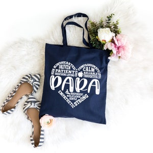 May include: A navy blue tote bag featuring a white heart-shaped graphic composed of words including "PAPA" and "KINDHEARTED," "DRIVEN," "PATIENT," "CALM," "RELIABLE," "THOUGHTFUL," "EMPATHETIC," and "STRONG," accompanied by a pair of striped flats.