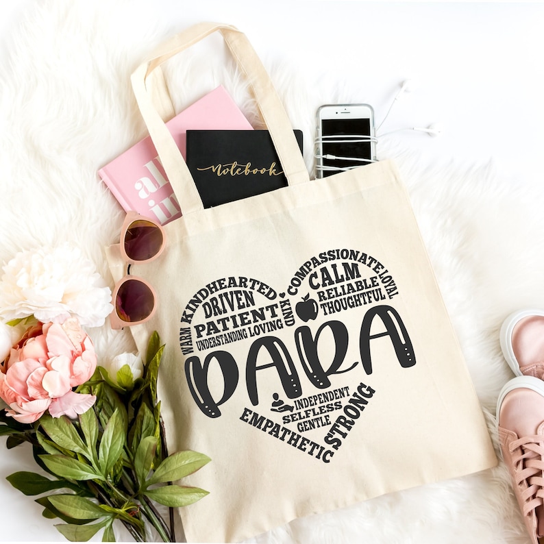 May include: A beige canvas tote bag featuring a black heart-shaped graphic composed of words including PARA, KINDHEARTED, DRIVEN, PATIENT, COMPASSIONATE, LOYAL, CALM, RELIABLE, THOUGHTFUL, INDEPENDENT, SELFLESS, GENTLE, EMPATHETIC, and STRONG with an apple icon.