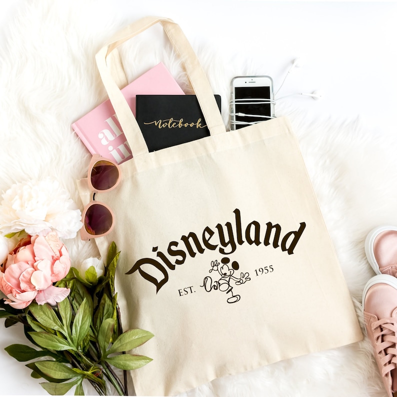 May include: A cream-colored canvas tote bag featuring the dark brown text Disneyland, the abbreviation EST., the year 1955, and a line-art illustration of Mickey Mouse. The bag contains a pink book, a black notebook, and a smartphone with white earbuds.