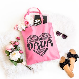 May include: A pink tote bag featuring a black heart-shaped graphic composed of words including PARA, KINDHEARTED, DRIVEN, PATIENT, COMPASSIONATE, CALM, RELIABLE, THOUGHTFUL, INDEPENDENT, SELFLESS, GENTLE, EMPATHETIC, and STRONG, with a small apple icon.