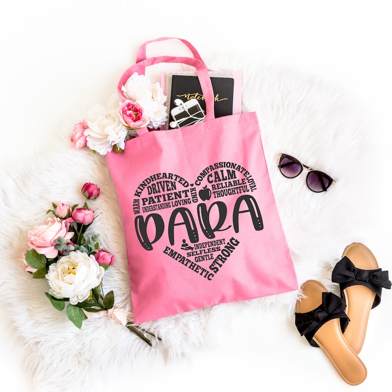 May include: A pink tote bag featuring a black heart-shaped graphic composed of words including PARA, KINDHEARTED, DRIVEN, PATIENT, COMPASSIONATE, CALM, RELIABLE, THOUGHTFUL, INDEPENDENT, SELFLESS, GENTLE, EMPATHETIC, and STRONG, with a small apple icon.