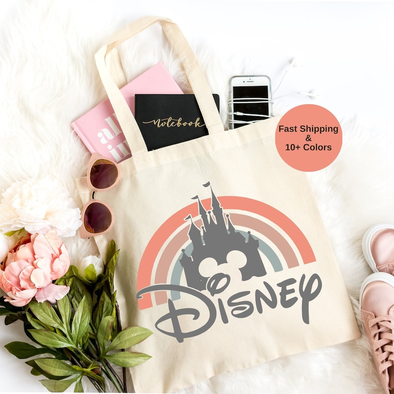 K&ouml;nnte beinhalten: Eine beige Canvas-Tragetasche mit einem Regenbogen und einer grauen Silhouette von Schloss Aschenputtel und dem Wort "DISNEY" in grauer Schrift.