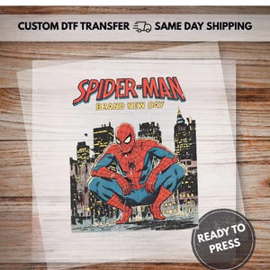 Puede incluir: Una hoja de transferencia DTF personalizada con un gráfico de Spider-Man agachado frente al horizonte de una ciudad. El texto dice SPIDER-MAN BRAND NEW DAY en la parte superior y READY TO PRESS en una insignia circular en la esquina inferior.