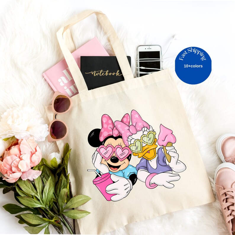 Op de afbeelding: Een beige canvas draagtas met een grafische print van Minnie Mouse en Daisy Duck met hartvormige zonnebrillen. Minnie houdt een roze drankje vast en Daisy een roze ijshoorntje. Beide figuren dragen roze strikken met stippen.