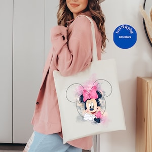 May include: A white canvas tote bag featuring a graphic of Minnie Mouse inside a black outline of Mickey Mouse ears. The design includes watercolor-style splashes of pink, purple, and grey behind the character. The bag has two shoulder straps.