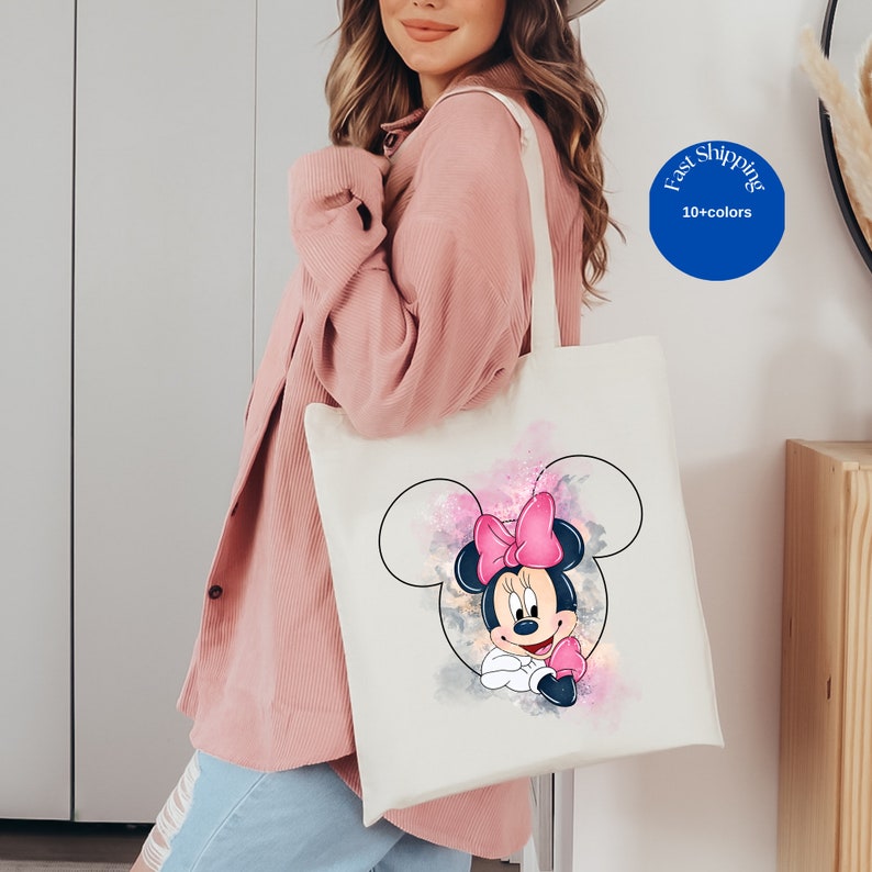 May include: A white canvas tote bag featuring a graphic of Minnie Mouse inside a black outline of Mickey Mouse ears. The design includes watercolor-style splashes of pink, purple, and grey behind the character. The bag has two shoulder straps.