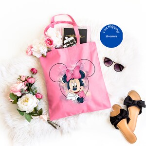 May include: A bright pink tote bag featuring a graphic print of Minnie Mouse with a pink bow, set against a watercolor-style background. The bag has two top handles and is displayed alongside white artificial flowers, black sunglasses, and sandals.