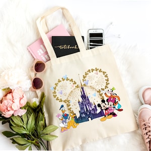 Puede incluir: Un bolso de tela color crema con un estampado gráfico de un castillo morado, fuegos artificiales dorados con purpurina en forma de orejas de Mickey Mouse y personajes de Disney como Mickey, Minnie, Donald, Daisy, Pluto y Goofy al frente.