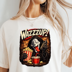 Pu&ograve; includere: Una t-shirt color crema con la grafica di una figura mascherata in una veste nera con cappuccio che tiene in mano un cellulare vintage, un secchiello per popcorn a righe rosse e bianche e un bicchiere rosso. Il testo WAZZZUP! &egrave; in lettere beige.