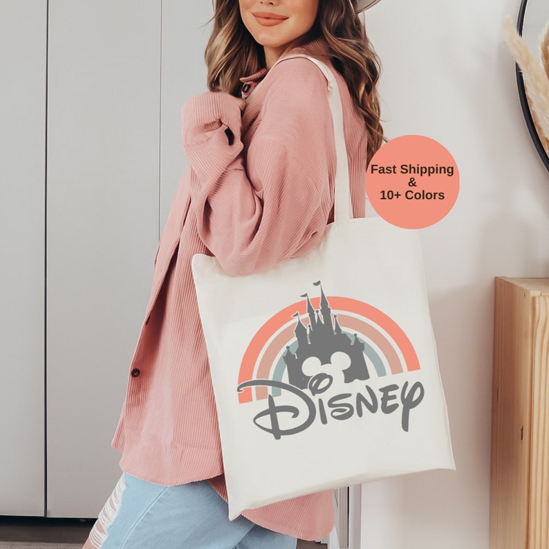 K&ouml;nnte beinhalten: Eine wei&szlig;e Canvas-Tragetasche mit einem Regenbogen und einer Silhouette von Schloss Aschenputtel mit dem Wort "DISNEY" in grauer Schrift.